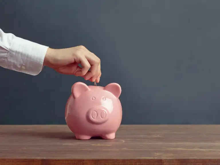 Mão inserindo uma moeda em um cofrinho de porco rosa sobre uma mesa de madeira, em um fundo cinza