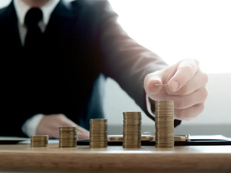 Mão de um profissional de terno adicionando uma moeda a uma das pilhas crescentes de moedas sobre uma mesa