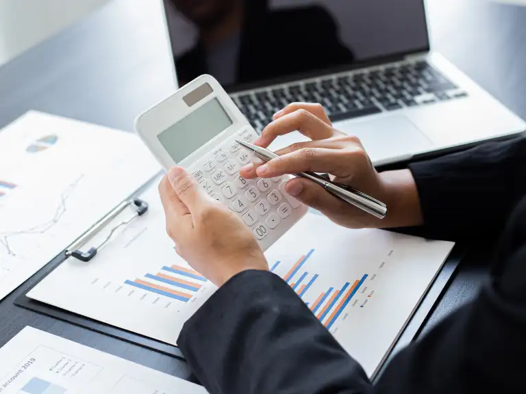 Pessoa usando uma calculadora e caneta sobre documentos financeiros e gráficos, com um laptop ao fundo