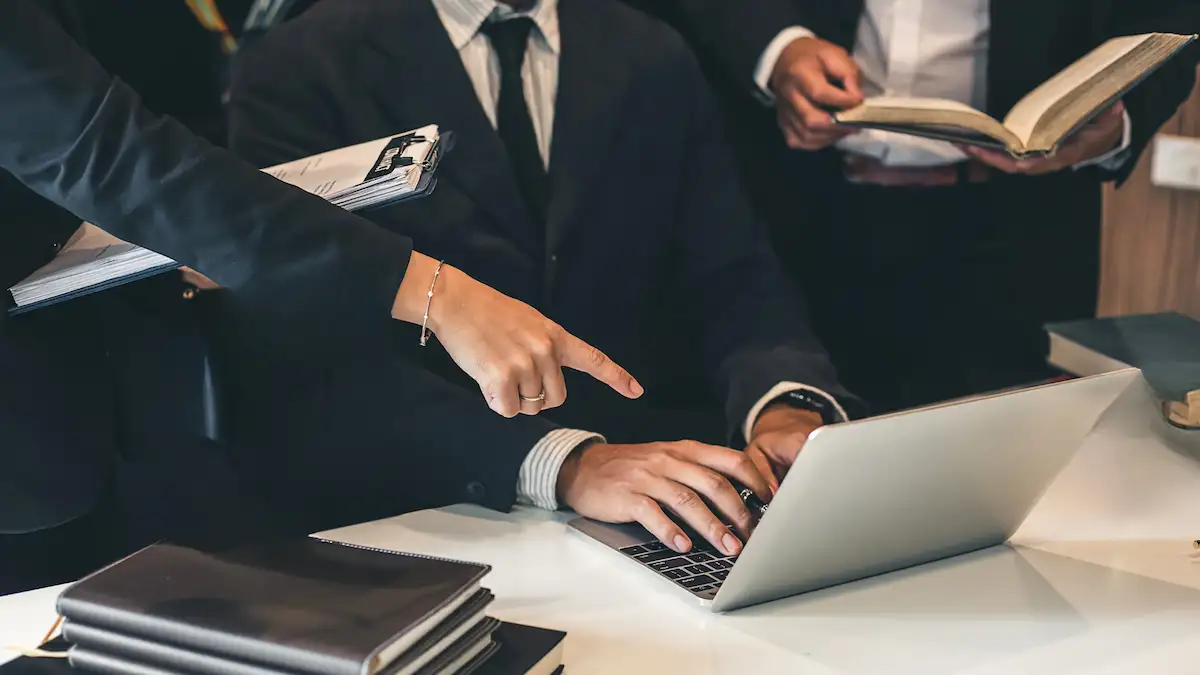Profissionais em trajes sociais colaboram em torno de um laptop, com uma pessoa apontando para a tela e outra segurando um livro